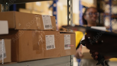 Young man using scanner on boxes barcodes tags in fulfillment center. Organized shelving, package labels and route planning support efficient distribution and shipment. Logistics hub. Camera B.
