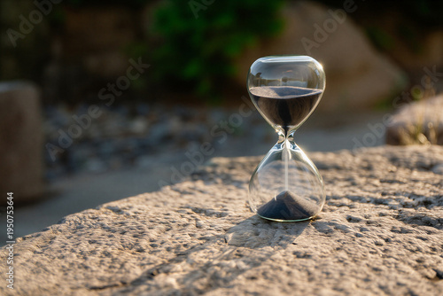 Hourglass with black sand on natural stone surface outdoors with blurred background for time and patience concept