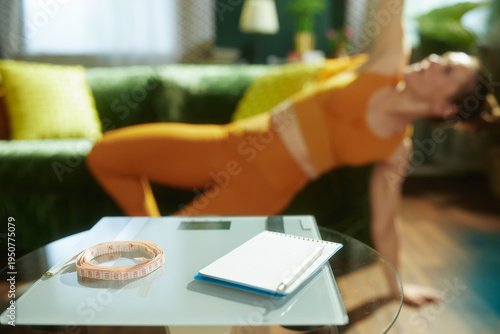Health tracking tools including a scale and measuring tape sit on a table while a woman in orange fitness gear performs a side plank in the sunlit background.