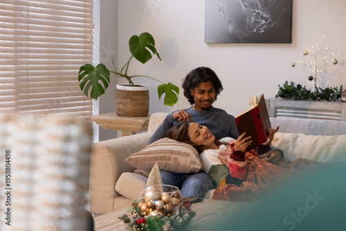 Diverse couple reclining on light-colored sofa at home, man reading red hardcover book