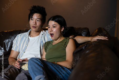 Asian couple sitting close on dark brown leather couch at home, holding popcorn bowl watching TV