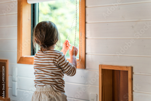Fotografie A mischievous girl's back as she plays by pulling on the cords of roller blinds and other blinds