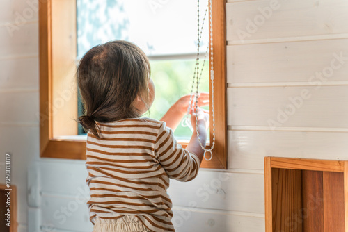 Fotografie A mischievous girl's back as she plays by pulling on the cords of roller blinds and other blinds