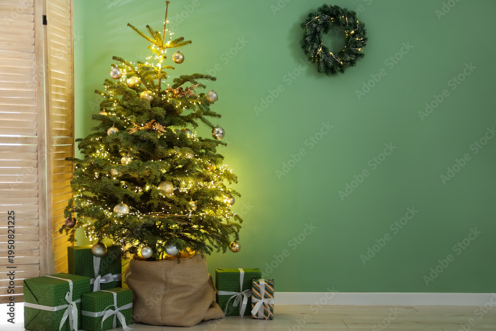 Fototapeta premium Beautiful fir tree with Christmas decor in pot, gift boxes and folding screen near green wall indoors. Space for text