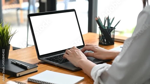 Person's hands typing on a laptop with a blank screen on a wooden desk.