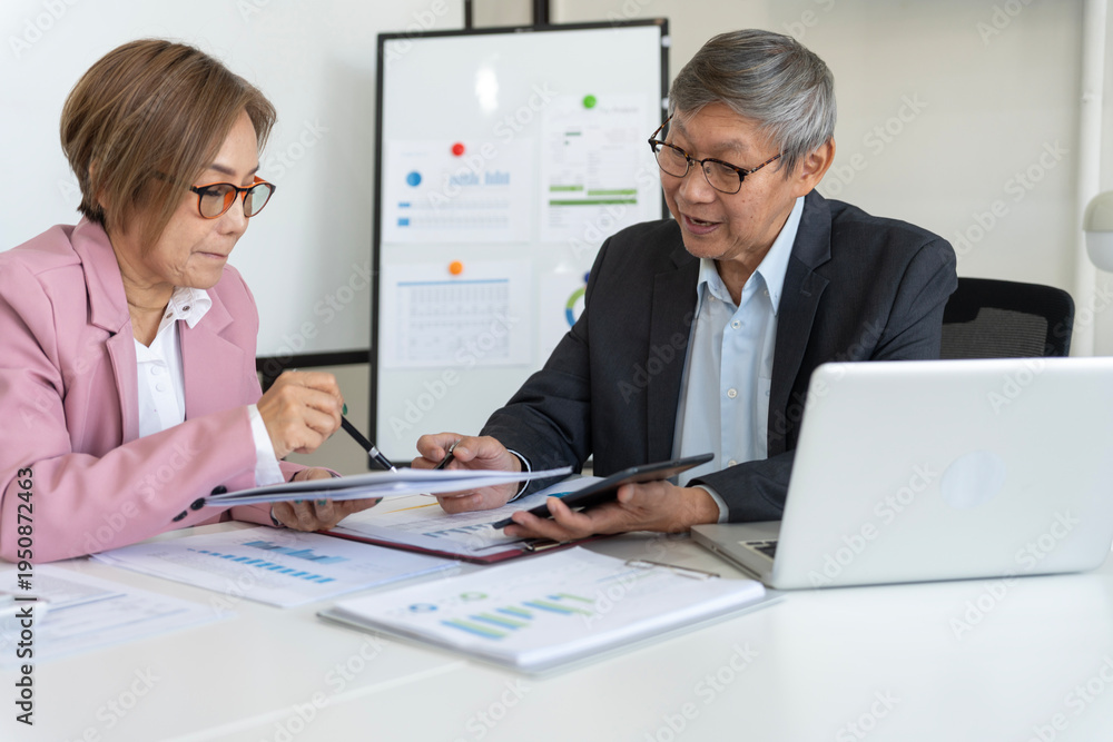 Fototapeta premium Two Senior Asian Business Professionals Reviewing Documents And Using Tablet Laptop At Office Meeting Table Whiteboard Background