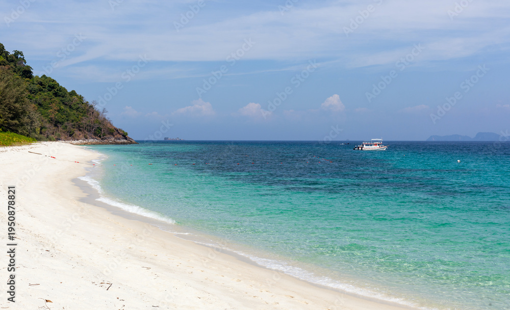 Fototapeta premium Tropical Koh Adang Beach in Thailand. White sand shoreline with turquoise water and clear blue sky. Boat in distance with lush green hills nearby.