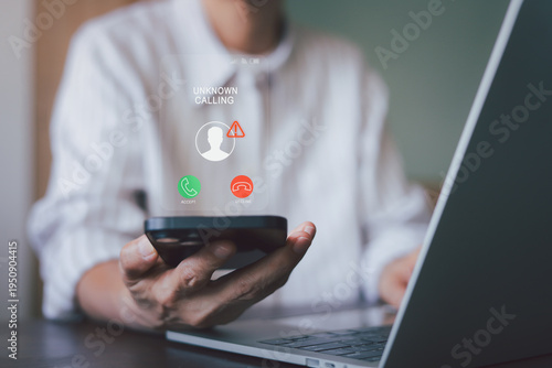 Businessman working on laptop with unknown call and warning icon on smartphone. Concept of cyber crime, phishing scam, and telemarketing fraud at workplace. Digital security and privacy protection.