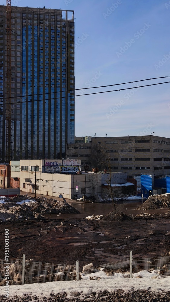 Fototapeta premium Urban construction site with exposed earth and partial snow in city. Tall skyscraper building under urban construction, rising above older city structures and power lines.