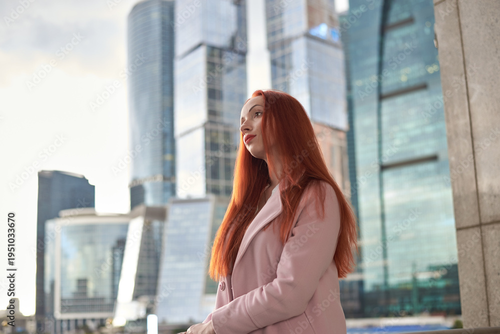 Fototapeta premium A red-haired woman stands in front of modern glass skyscrapers. She is wearing a light pink coat and is looking directly at the camera with a calm, composed expression.