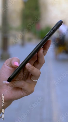 Closeup of female hand hold and touch screen smartphone while horses and carriages pass of background. Asian woman using cell phone in Seville street. Girl messaging on smart-phone in Spain