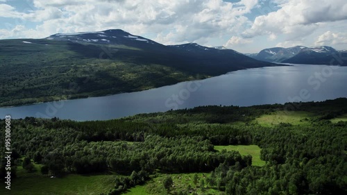 Drone rising up at Gjevilvatnet lake in Oppdal, Norway. Views of the Trollheimen mountain range and green meadows and forests fill the landscape on a sunny summer day.
