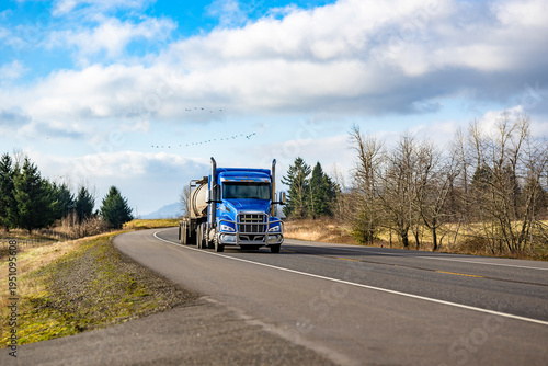 Bright blue day cab big rig semi truck tractor transporting liquid cargo in tank semi trailer driving on the flat highway road at sunny day