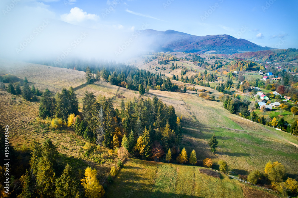 Obraz premium Picturesque village nestled in rolling hills, bathed in autumn colors with patches of green and yellow fields. Wisps of fog drift through landscape, adding dreamy atmosphere to serene rural scene.