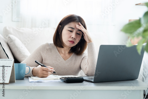 Stressed Asian woman using calculator and laptop to manage debt at home. Concept of financial crisis, unpaid bills, bankruptcy, household expenses, business failure, and money problems stress