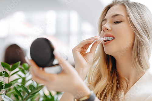 Young woman inserting clear dental aligner while checking her smile in a mirror at home