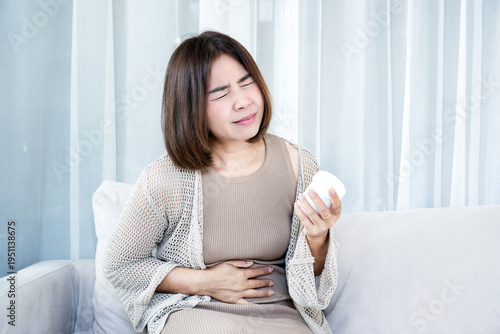 Asian woman suffering from severe stomach ache and abdominal pain while holding medicine bottle on sofa at home. Concept of gastritis, food poisoning, indigestion, and feminine healthcare