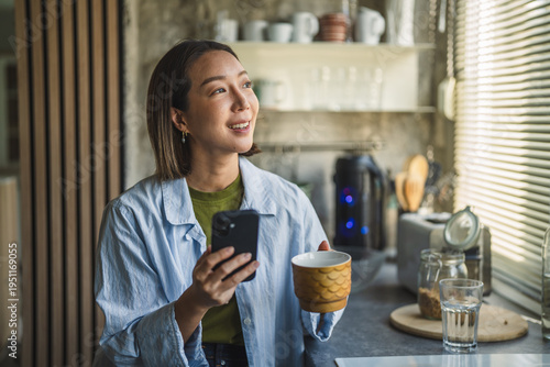 Woman smiling using smart phone and drinking coffee in kitchen
