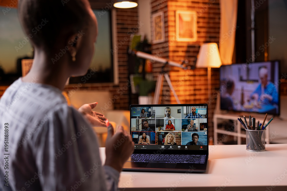 Fototapeta premium Young black woman working remotely from cozy living room during sunset, attending online video conference. Female entrepreneur having virtual discussion with diverse coworkers on laptop.
