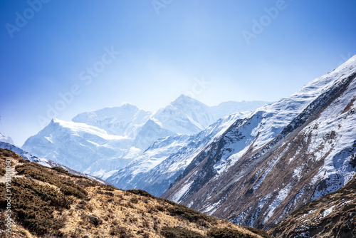 Schilderij op canvas Breathtaking view of snow-covered Himalayan peaks rising above a dramatic alpine valley, with a winding trail cutting through dry grassland in the foreground