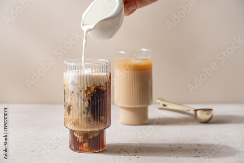 Milk pouring into iced black coffee in glass on light background with copy space, creamy cold drink and iced latte concept