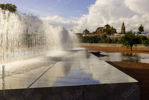 A person stands near a fountain with the Mezquita-Cathedral of Córdoba in the background under a cloudy sky.
