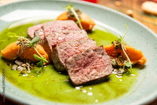 Juicy beef steak slices on rich green sauce with carrot puree and microgreens, beautifully plated on ceramic plate, rustic wooden table