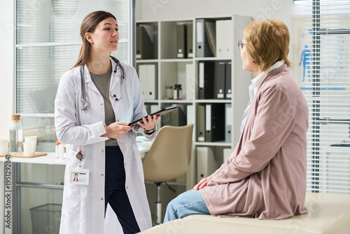 Konstfotografi Young female doctor holding digital tablet speaking with senior woman sitting on
