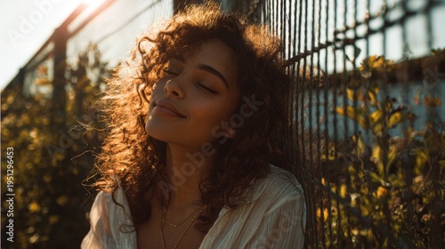 Young woman smiling with closed eyes against metal fence at sunset