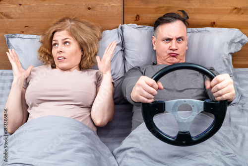 Fotografia Funny couple in bed: man grips a steering wheel pretending to drive, while woman throws hands up in disbelief