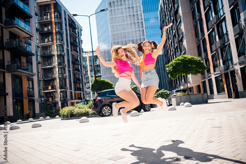 Naklejka premium Two young girlfriends jump in the city street wearing bright outfits enjoying sunny day urban fashion and friendship while exploring modern city life and feeling joyful energetic and carefree together