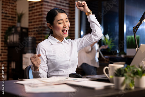 Bild auf Leinwand Happy asian entrepreneur celebrates a win at work, raising her arms in excitement while looking at her laptop