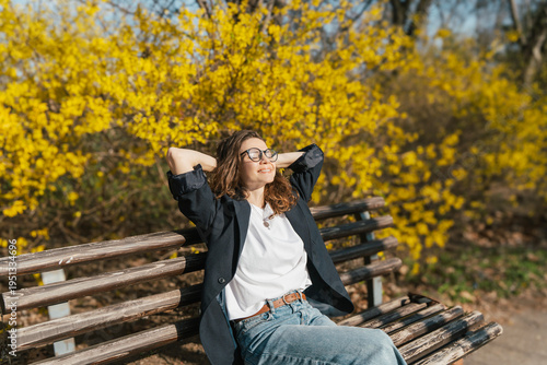 Relaxed young curly woman in glasses enjoying spring sun on a city bench under blooming yellow forsythia bushes