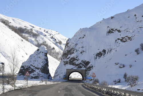 a paved mountain road leading toward a stone-arched tunnel carved through a steep snow-covered rocky