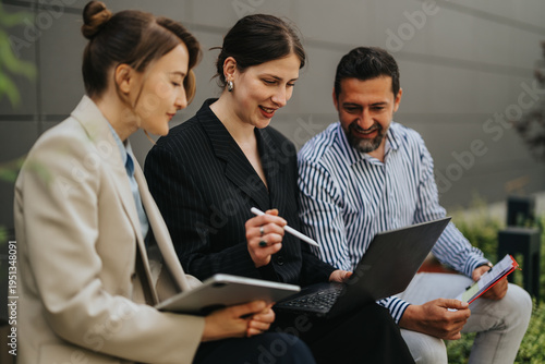 Three professionals collaborate while reviewing a laptop and tablet during an outdoor meeting. A small team discusses work and shares ideas around a laptop and digital tablet.