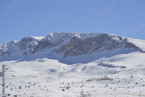 a massive snow-covered mountain range stretching across the frame under a clear pale blue sky the rugged cliffs and steep rocky faces are partially dusted with white snow