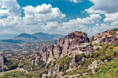 View of towering rock formations crowned with monasteries stand sentinel under a sky ablaze with azure and cottony clouds, Kalabaka, Trikala, Greece.
