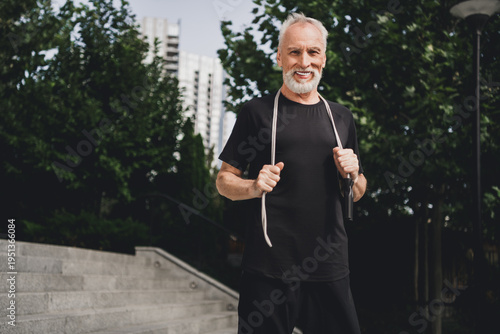 Senior bearded man smiles while jumping rope outdoors in a city park for fitness lifestyle and active wellness