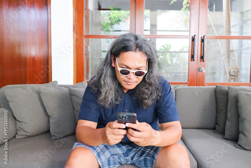 Obraz na płótnie Indonesian man sitting down on a couch and read a message on a mobile phone