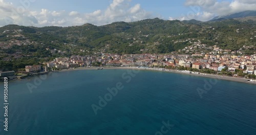 Panoramic aerial shot of a Mediterranean town sprawling along a sandy beach. Lush green mountains form a dramatic backdrop under a soft blue sky with clouds. It is Sapri, a town in Campania, Italy.