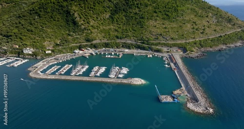 Aerial view of a long stone breakwater protecting a serene harbor filled with white boats. The deep blue sea contrasts with the rugged coastline. It's the Sapri port, in Campania, Southern Italy.