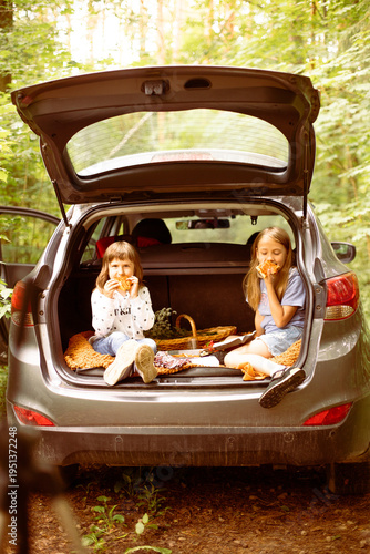 Sister girls are sitting in the trunk of the car and eating pizza. Picnic in the forest in the car in the summer