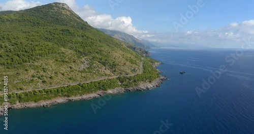 Aerial view of a winding coastal road carved into a steep, lush green mountain. The rocky shoreline meets the deep blue sea under a bright sky, highlighting a dramatic and wild Mediterranean landscape