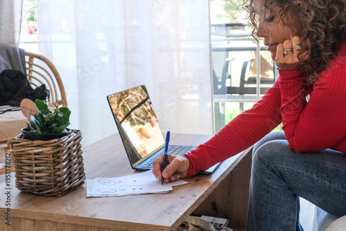 Focused adult woman working on laptop while writing notes on paper at home sofa, managing tasks and planning schedule. Professional freelancer organizing online business and productivity workflow in c