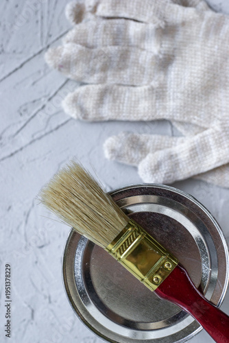 paint can sits on textured gray surface with brush resting on top. next to them, pair gloves is placed ready for painting task. tools are prepared for use. closeup.