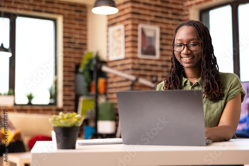 Φωτογραφία African american woman smiling brightly at device screen, reading online articles in brick wall apartment