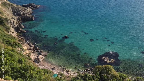 Aerial view of a rocky coastline with clear turquoise water. The beach is small and surrounded by green vegetation. Sunlight reflects on the water surface.