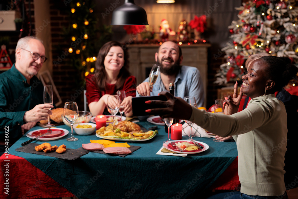Fototapeta premium People taking pictures at dinner table, making memories of christmas eve festivity gathered around at home. Diverse persons having fun with wine and food, taking photos with smartphone device.