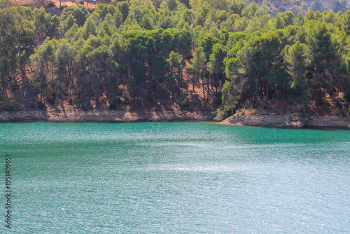 A scenic view along the banks of the El Chorro reservoir in Malaga, Spain. Vibrant green trees grow from the arid, sand colored earth, creating a striking contrast with the deep blue water of the lake