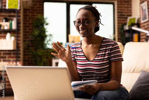 Quadro em tela Smiling black woman with pen in hand, waves at device screen, attending online business meeting from home
